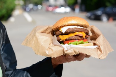 Photo of Woman holding delicious burger in paper wrap on city street, closeup