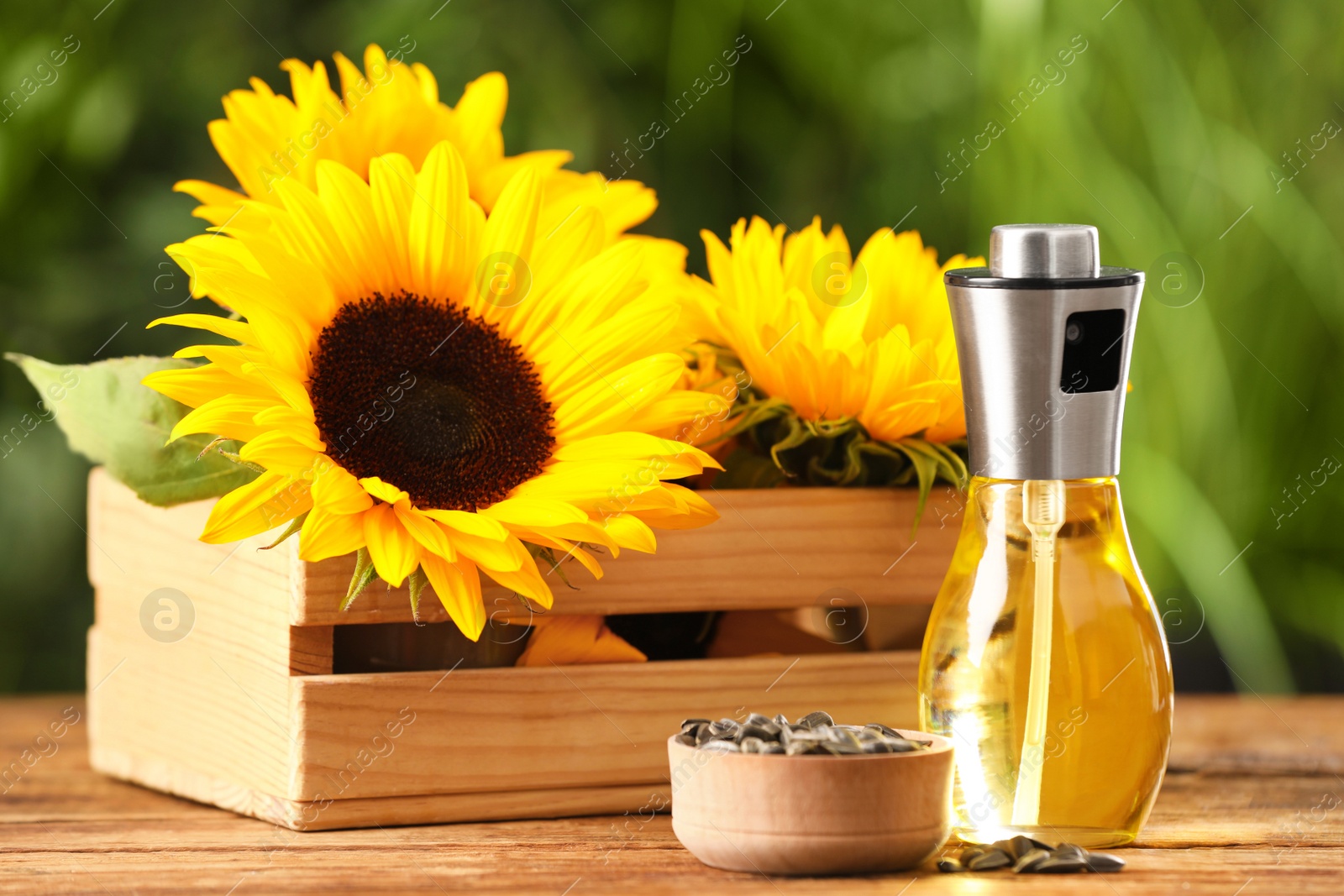 Photo of Spray bottle of cooking oil, sunflower seeds and crate with flowers on wooden table against blurred background