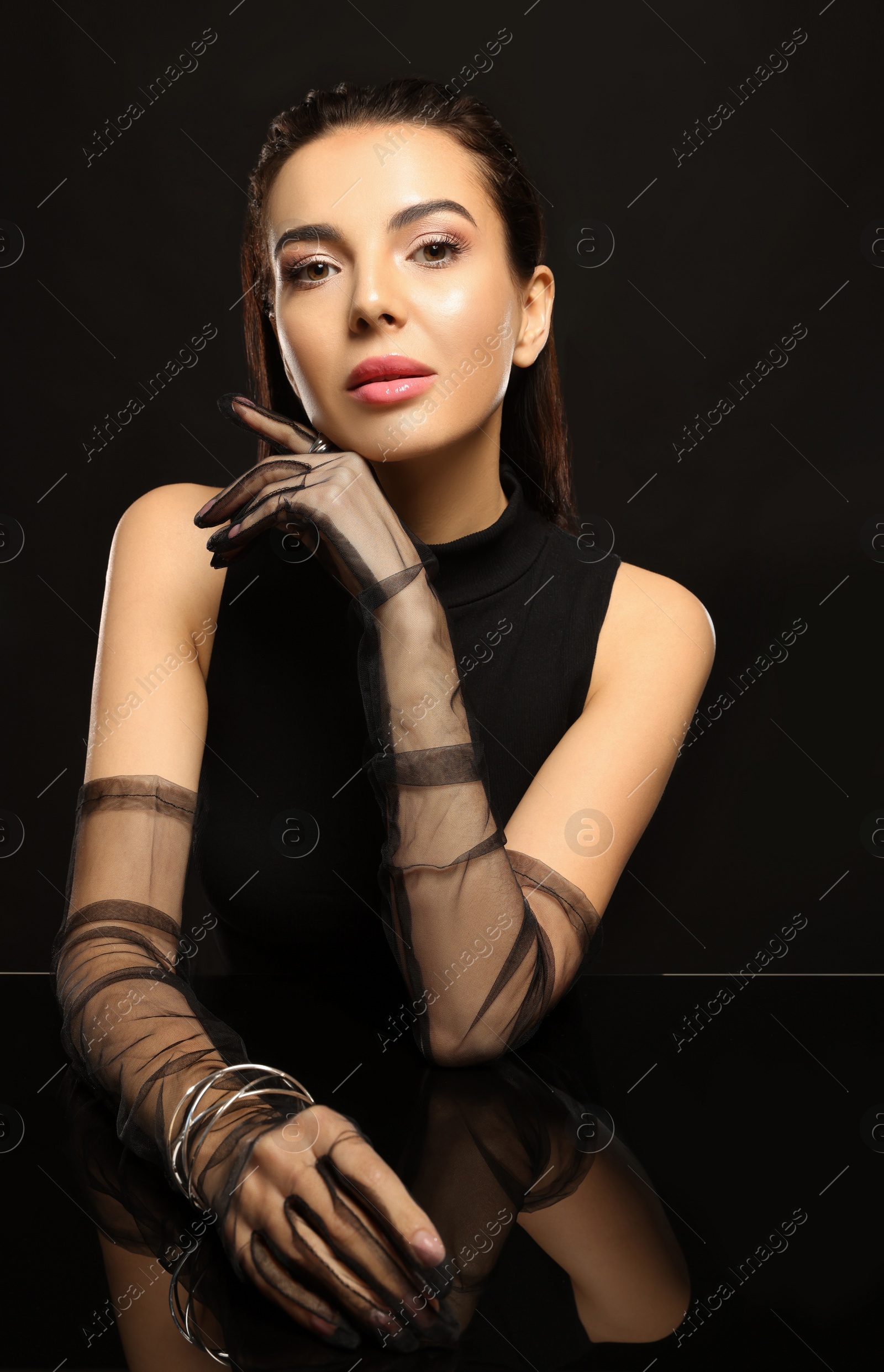 Photo of Beautiful young woman in evening gloves at black glass table against dark background
