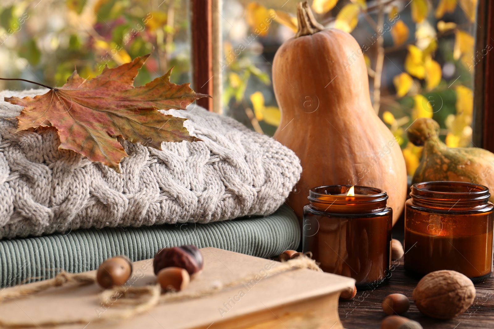 Photo of Burning scented candles, warm sweaters, book and pumpkins on wooden table near window. Autumn coziness