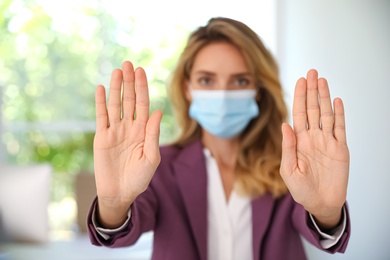 Woman in protective face mask showing stop gesture indoors, focus on hands. Prevent spreading of coronavirus