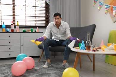 Photo of Tired man with bottle of detergent and rag in messy room after party