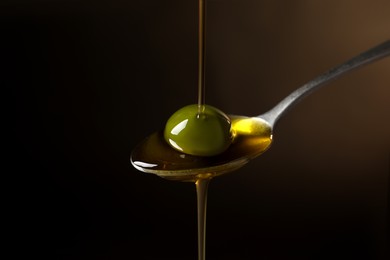 Photo of Pouring oil into spoon with ripe olive against dark background, closeup