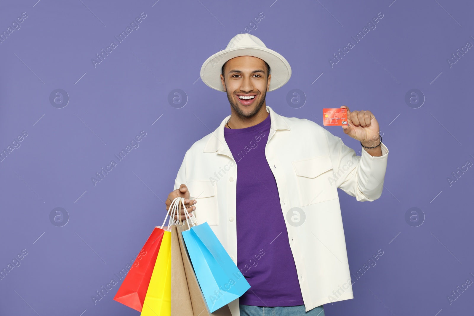 Photo of Happy African American man in hat with shopping bags and credit card on purple background