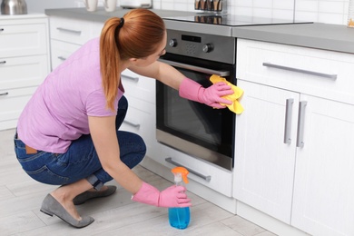 Woman cleaning modern oven with rag and detergent in kitchen