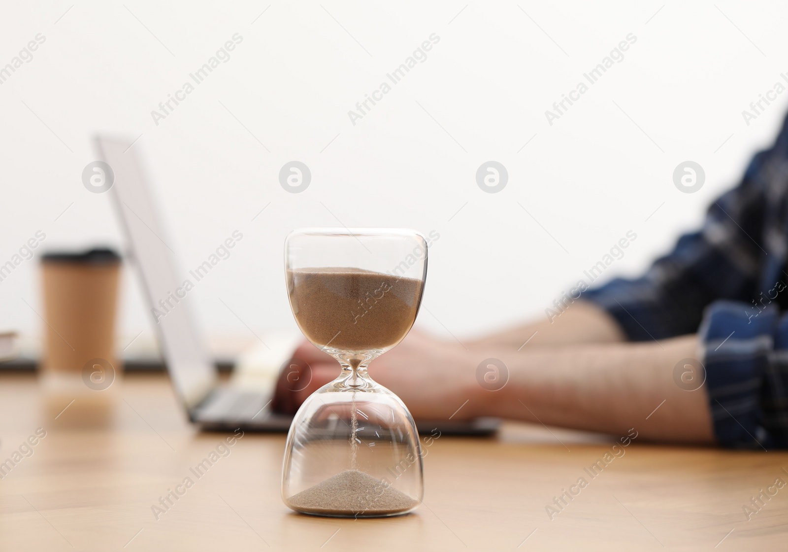 Photo of Hourglass with flowing sand on desk. Man using laptop indoors, selective focus