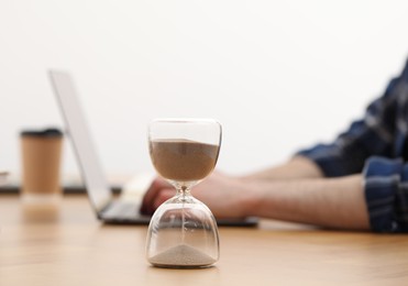 Hourglass with flowing sand on desk. Man using laptop indoors, selective focus