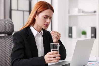 Photo of Woman with pill and glass of water suffering from headache at table in office