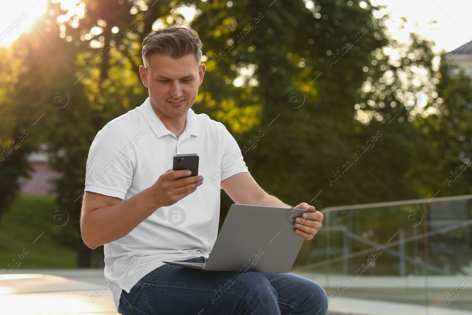 Photo of Handsome man using smartphone and laptop on bench outdoors. Space for text