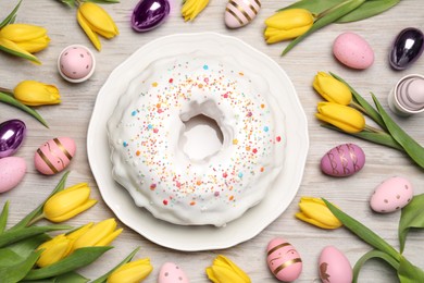 Photo of Easter cake with sprinkles, painted eggs and tulips on white wooden table, flat lay