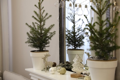 Photo of Little fir trees and Christmas decorations on shelf near mirror in room. Stylish interior design