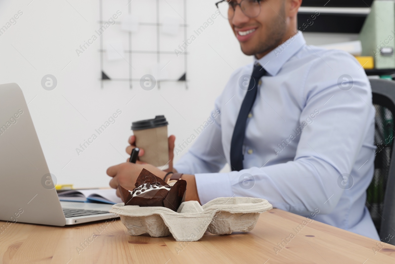 Photo of Happy young intern with paper cup of hot drink working on laptop at table in modern office, focus on muffin