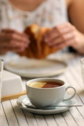 Photo of Woman eating croissant at table, focus on cup of aromatic tea with lemon