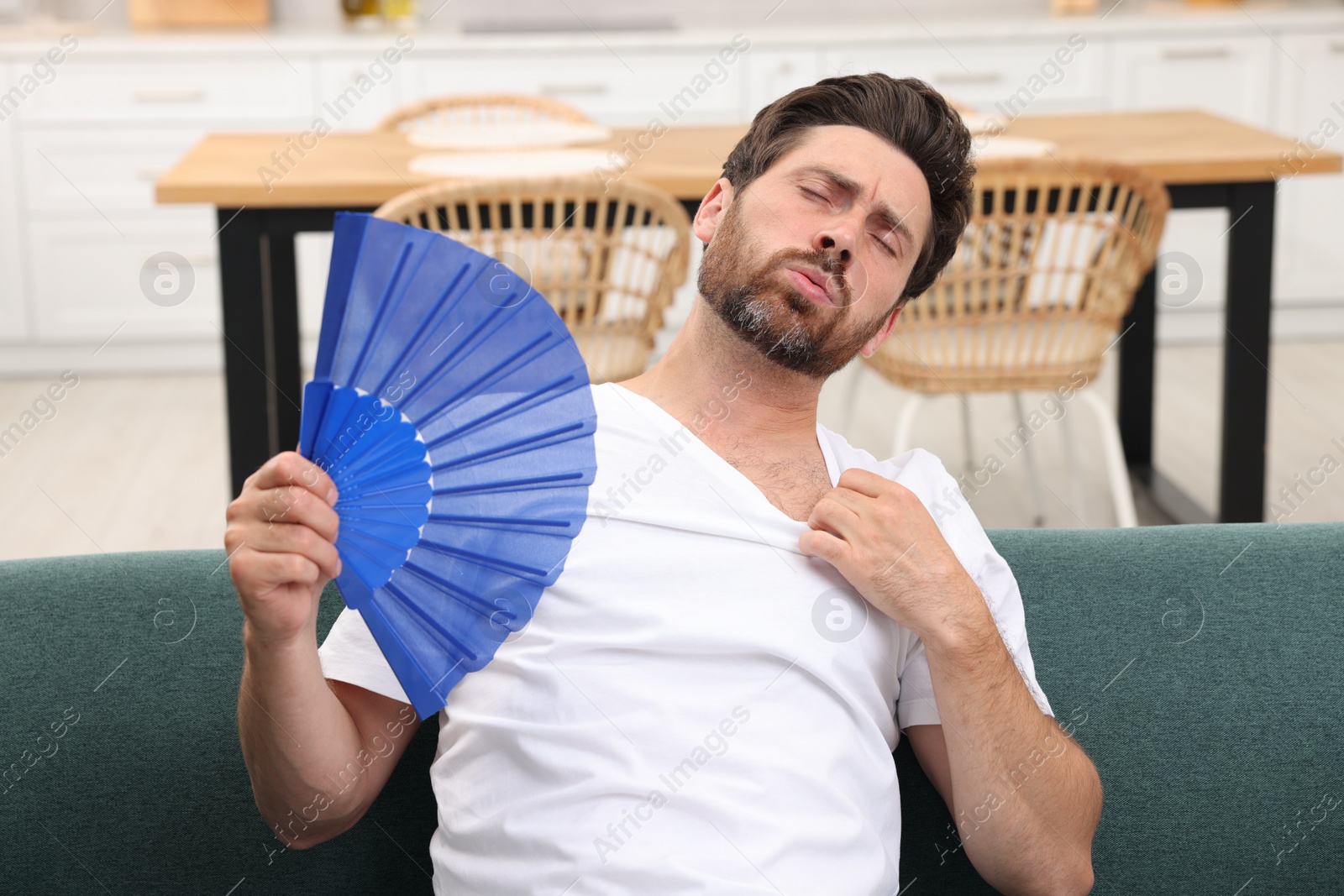 Photo of Bearded man waving blue hand fan to cool himself on sofa at home