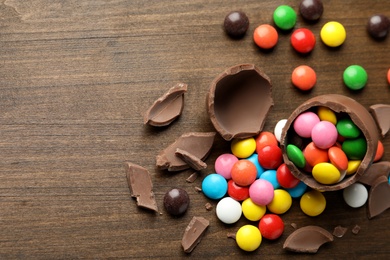 Photo of Broken chocolate egg and colorful candies on wooden table, flat lay. Space for text