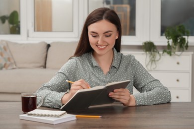 Happy young woman writing in notebook at wooden table indoors