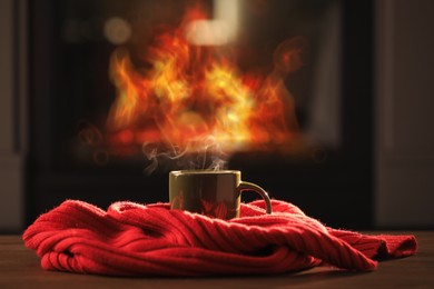 Photo of Cup of hot drink and red knitted scarf on wooden table near fireplace at home. Cozy atmosphere