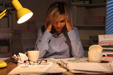 Overwhelmed woman surrounded by documents and dirty cups at table in office at night