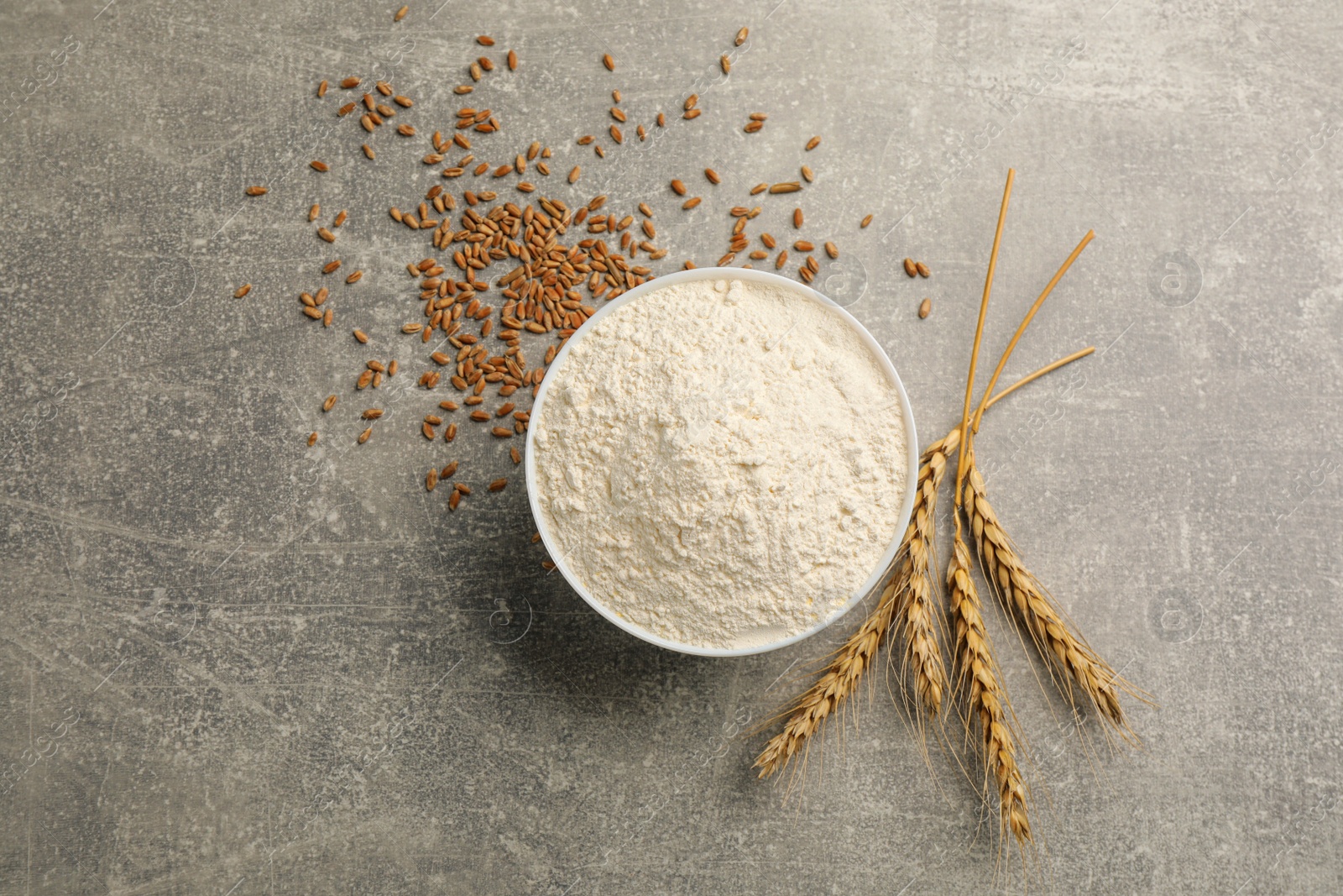 Photo of Bowl of flour, wheat ears and kernels on light grey table, flat lay