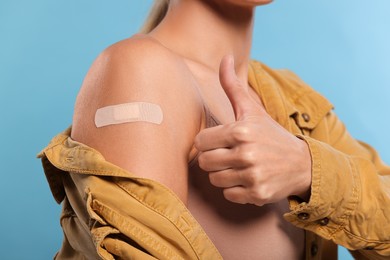 Photo of Woman with adhesive bandage on arm after vaccination showing thumb up on light blue background, closeup