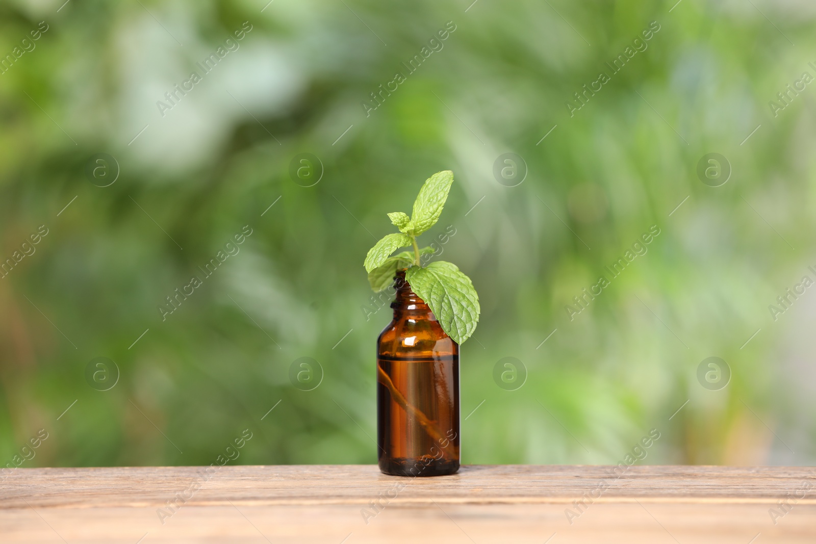 Photo of Bottle with essential oil and mint on wooden table against blurred green background