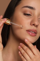 Photo of Beautiful woman applying essential oil onto face on brown background, closeup