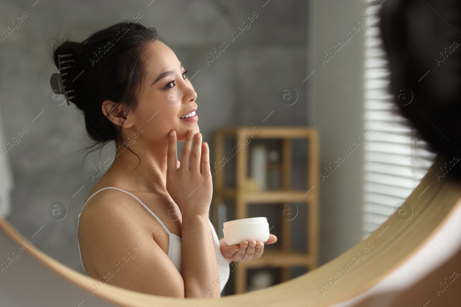 Photo of Happy woman applying face cream near mirror at home