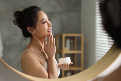 Happy woman applying face cream near mirror at home