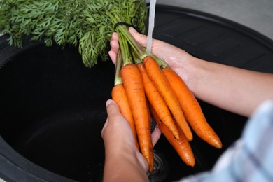 Photo of Woman washing ripe carrots with running water in sink, closeup