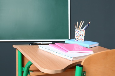 Wooden school desk with stationery near blackboard on grey wall