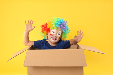 Photo of Little boy in clown wig sitting inside of cardboard box on yellow background. April fool's day