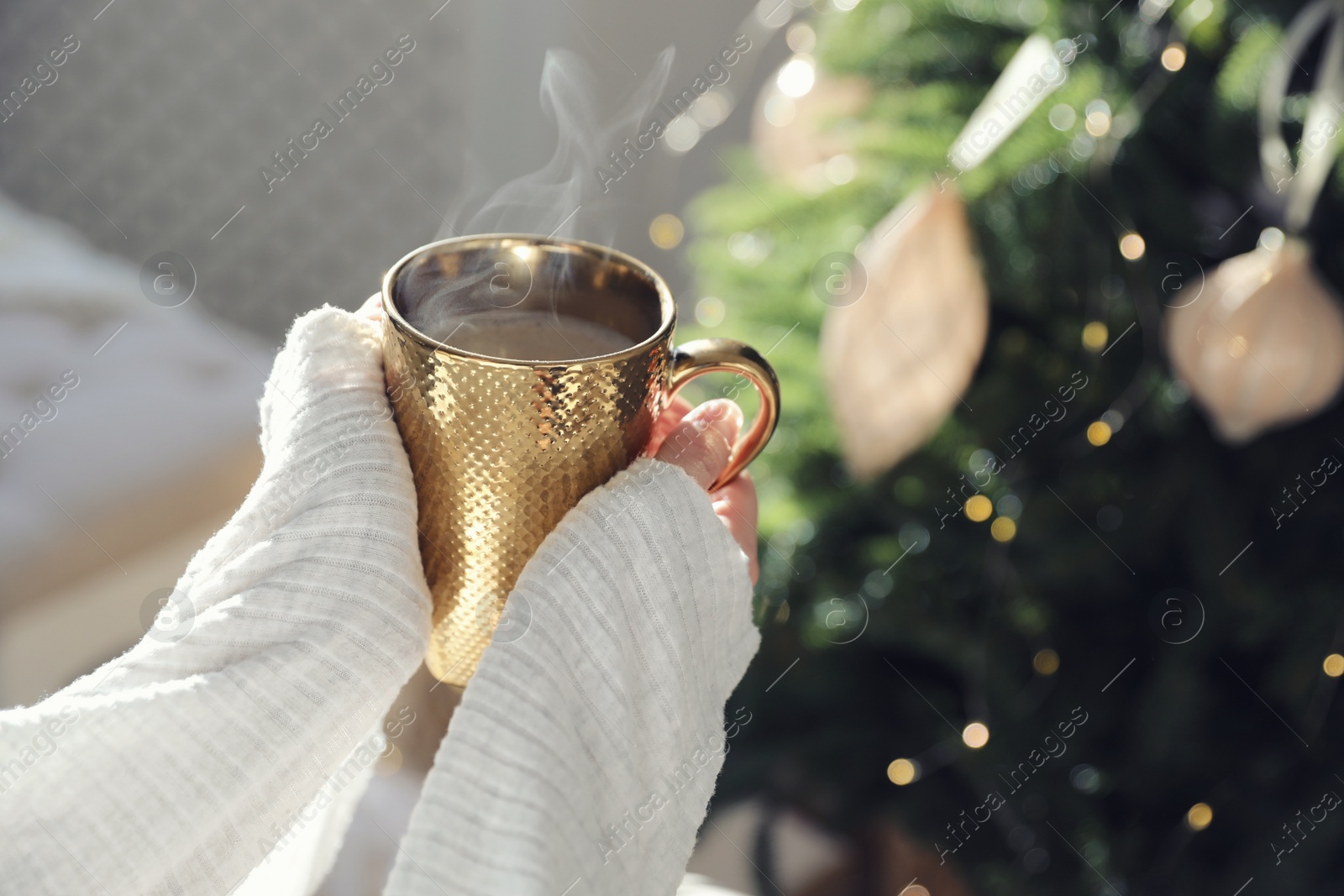 Photo of Woman with golden cup of hot drink near Christmas tree at home, closeup