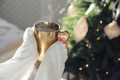 Photo of Woman with golden cup of hot drink near Christmas tree at home, closeup
