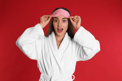 Photo of Young woman in bathrobe and eye sleeping mask on red background