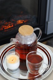 Teapot, cup of aromatic tea and burning candle on white table near fireplace indoors