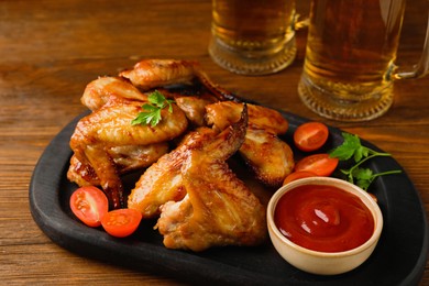 Mugs with beer, delicious baked chicken wings and sauce on wooden table