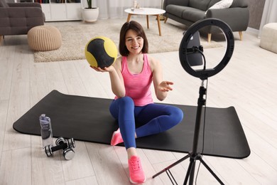 Happy sports blogger holding medicine ball while streaming online fitness lesson with smartphone at home