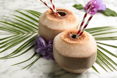 Photo of Fresh coconuts with drinking straws and flowers on white marble table, closeup