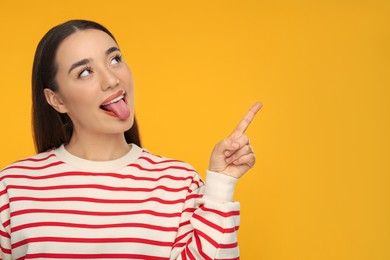 Photo of Happy woman showing her tongue and pointing at something on orange background. Space for text
