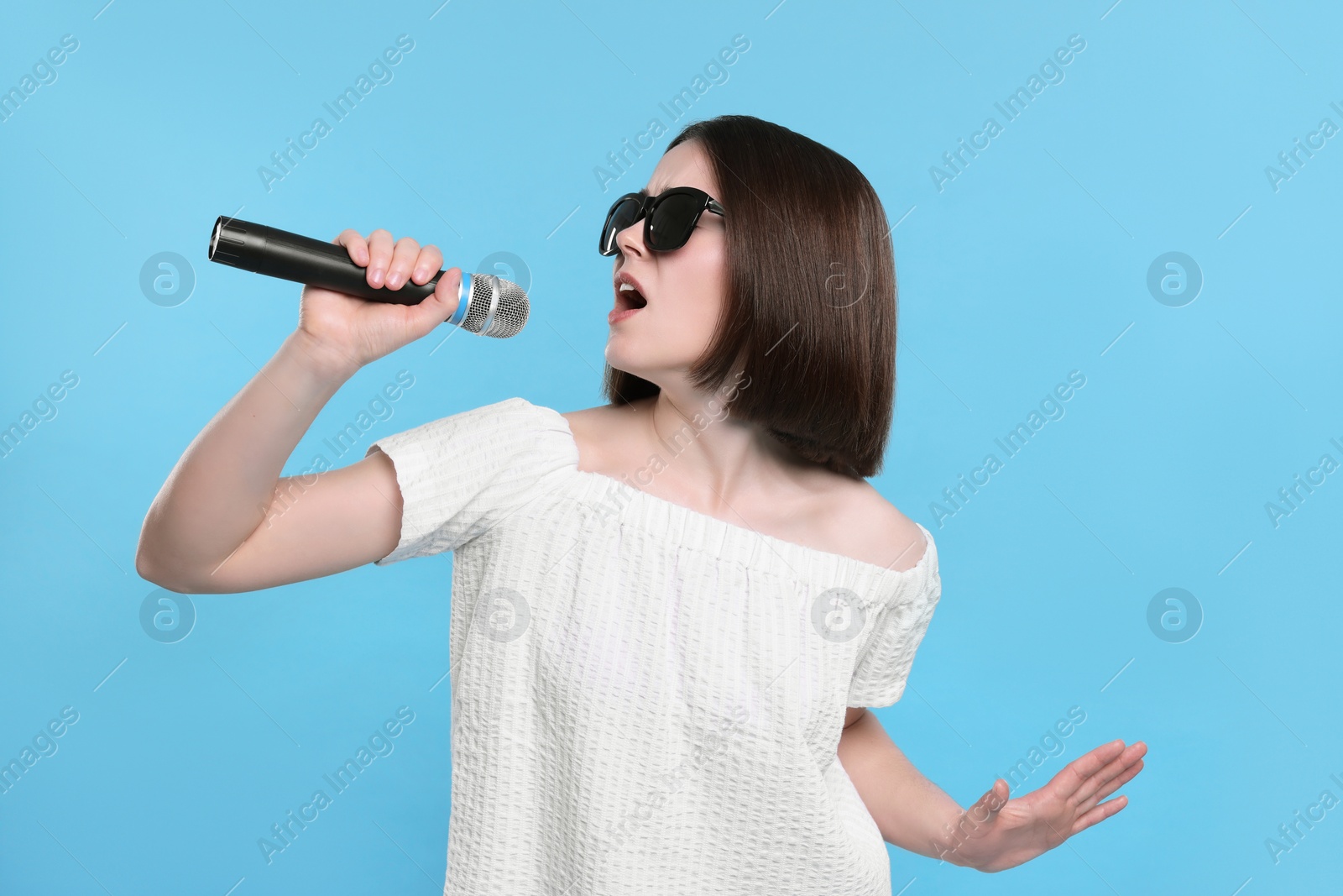 Photo of Beautiful young woman with sunglasses and microphone singing on light blue background