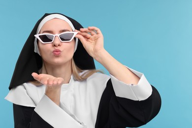 Woman in nun habit and sunglasses blowing kiss against light blue background. Space for text