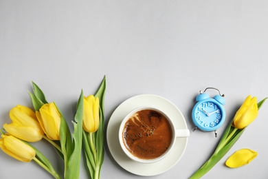 Aromatic morning coffee, alarm clock and beautiful flowers on light grey background, flat lay. Space for text