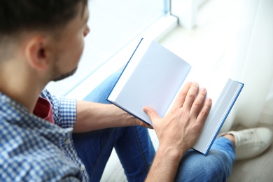 Photo of Handsome man reading book near window indoors, above view