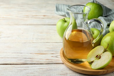 Photo of Jug of tasty juice and fresh ripe green apples on white wooden table, space for text