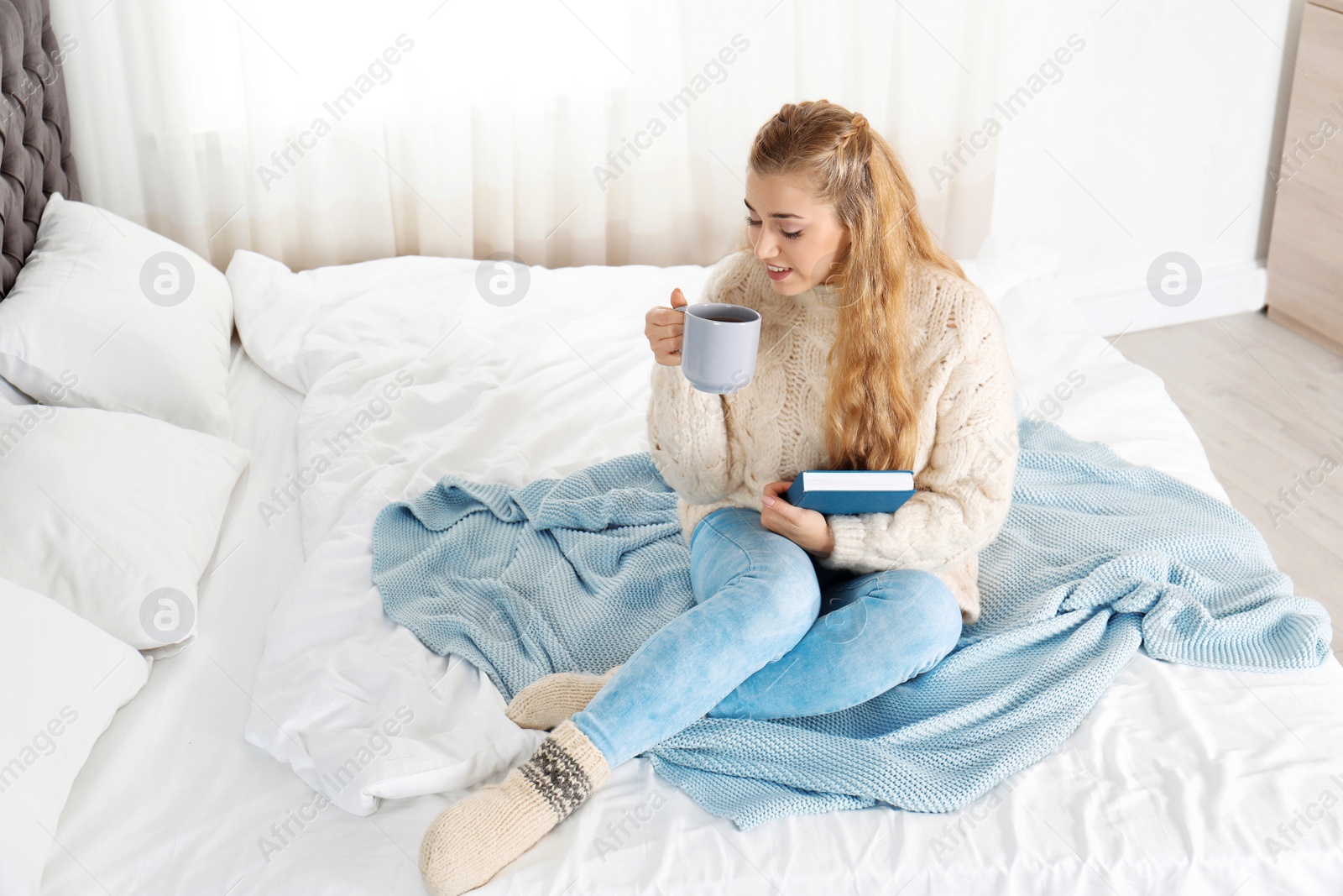 Photo of Attractive young woman in cozy warm sweater with cup of hot drink and book sitting on bed at home