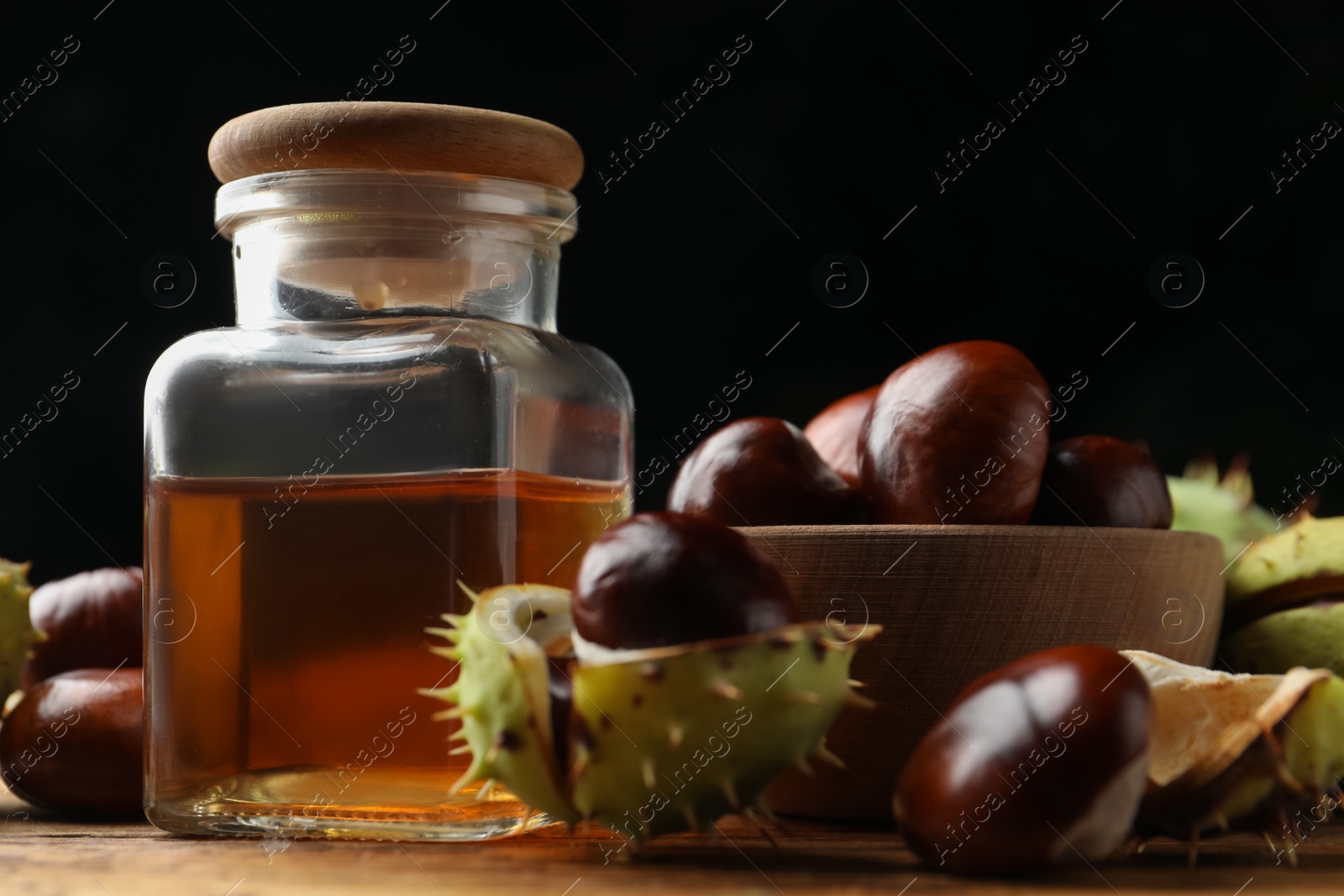 Photo of Chestnuts and jar of essential oil on wooden table against black background, closeup