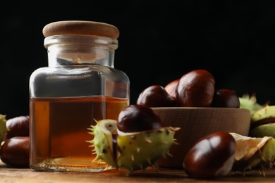 Chestnuts and jar of essential oil on wooden table against black background, closeup