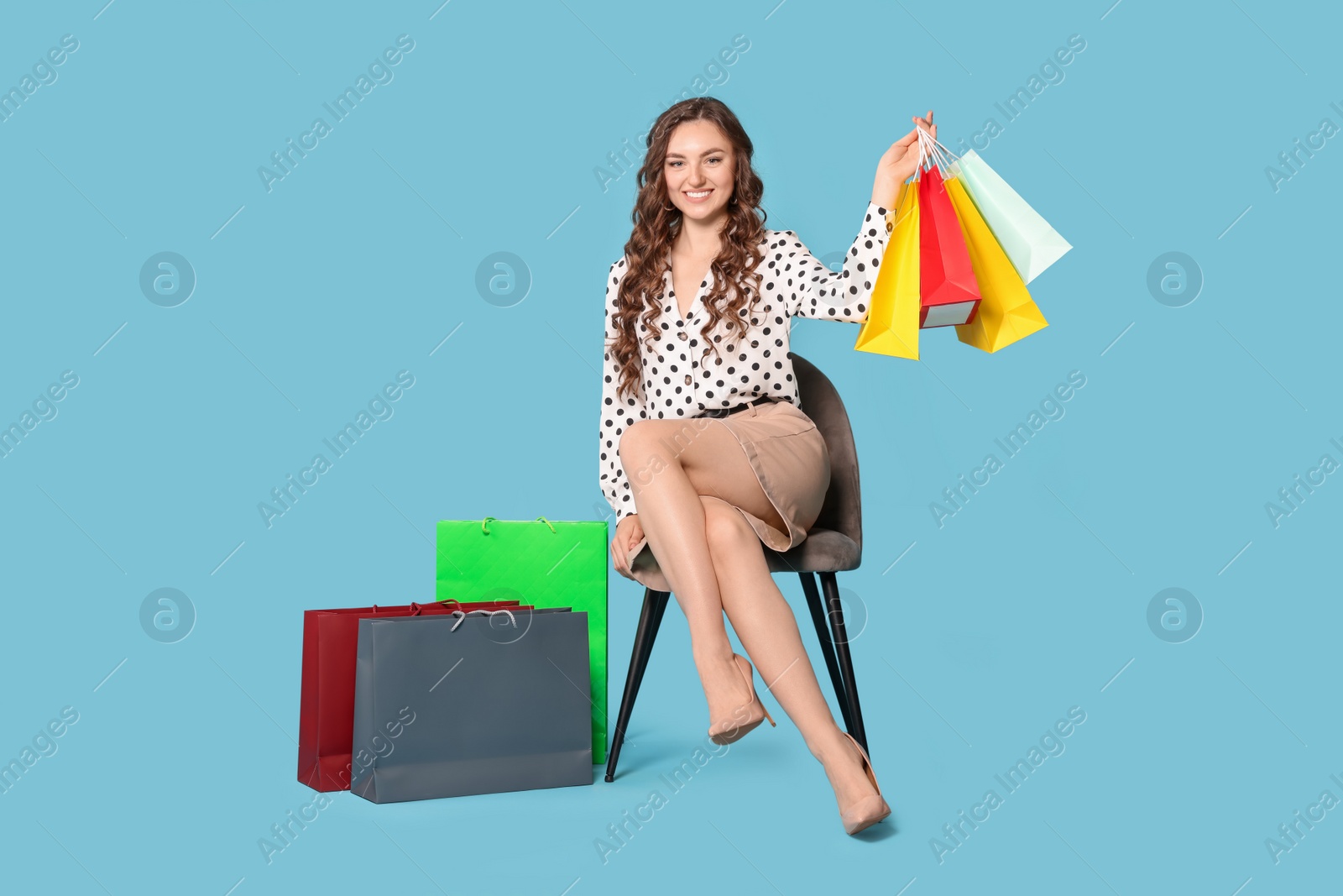 Photo of Happy woman holding colorful shopping bags on chair against light blue background