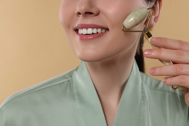 Young woman massaging her face with jade roller on beige background, closeup