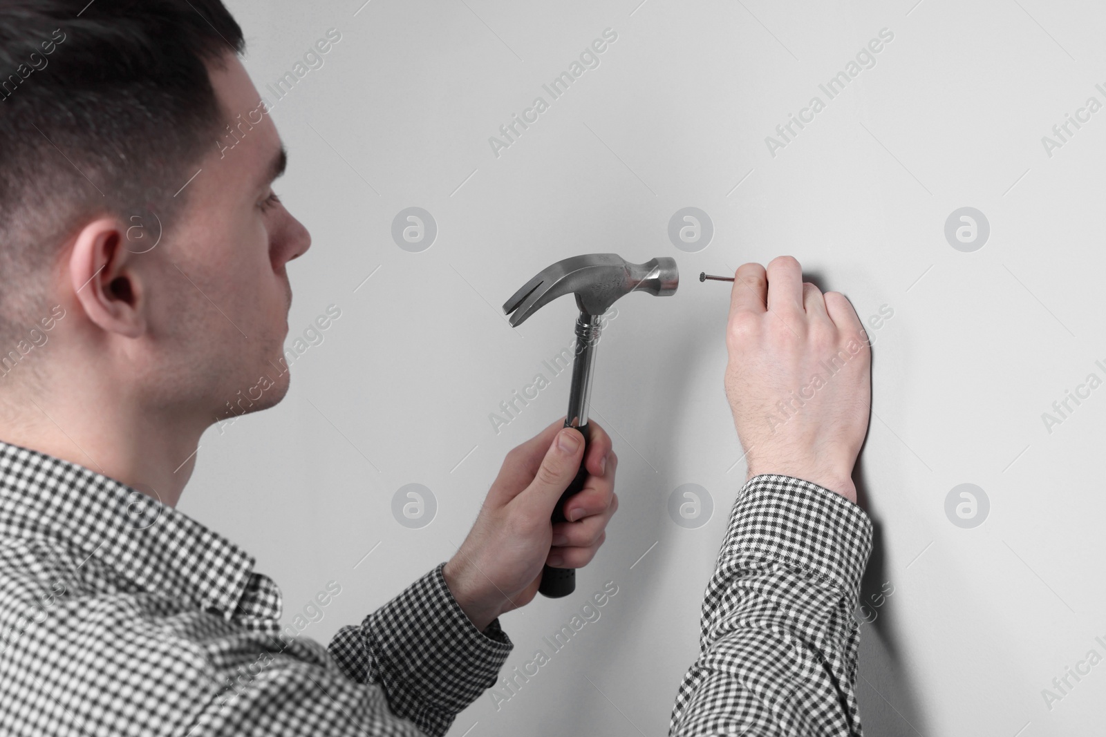 Photo of Young man hammering nail into white wall indoors